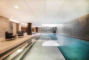 Une piscine intérieure moderne dotée d’une paroi en verre transparent, d’un espace détente avec des sièges et d’un plafond haut avec des luminaires encastrés.