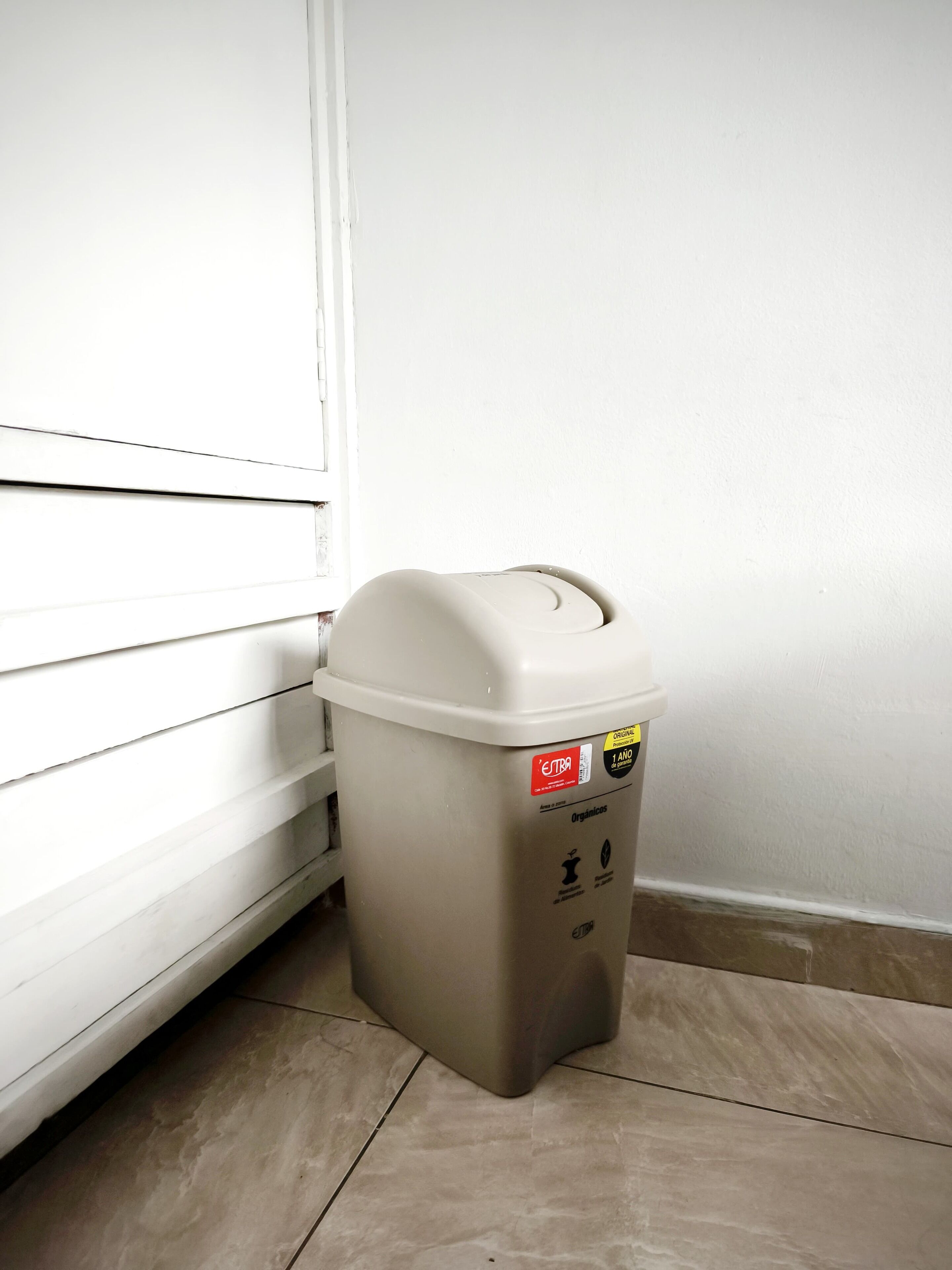 A beige trash can with a lid, placed against a white wall and wooden floor.