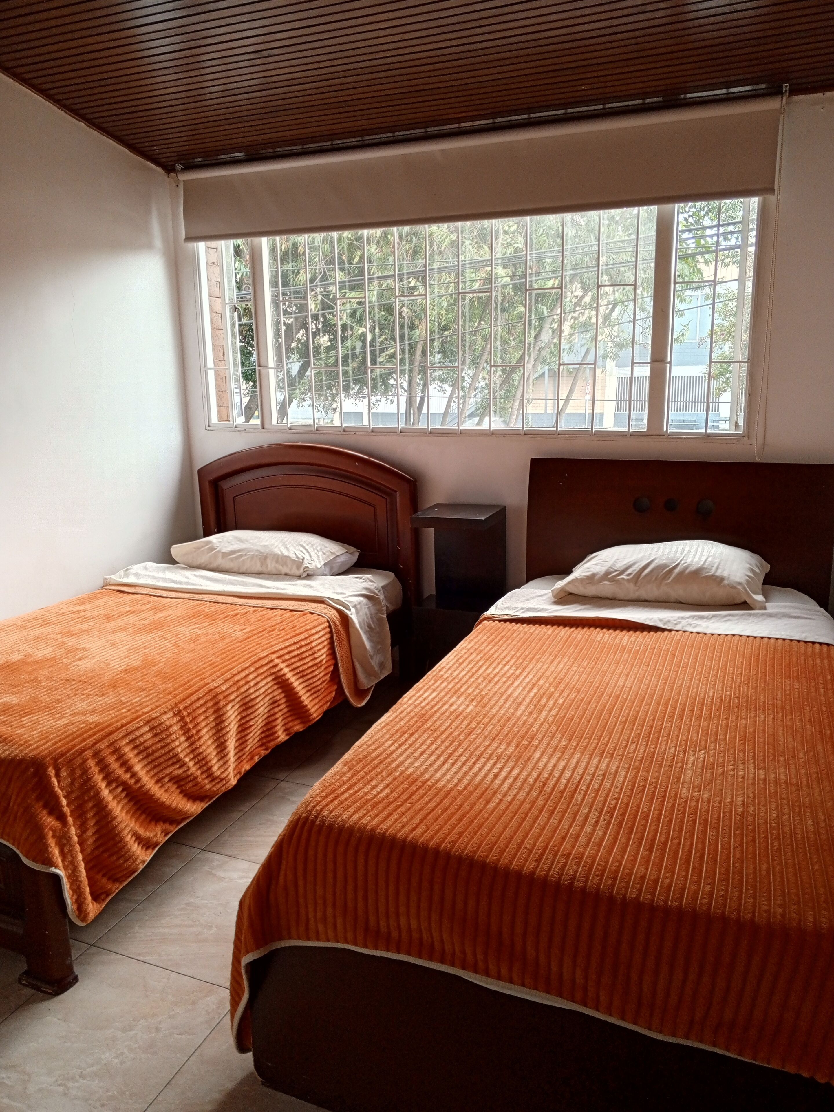 A room with two single beds, each with an orange bedspread and white pillows. There is a wooden headboard on each bed, and a small bedside table with a lamp. A window with metal bars is visible, showing greenery outside.
