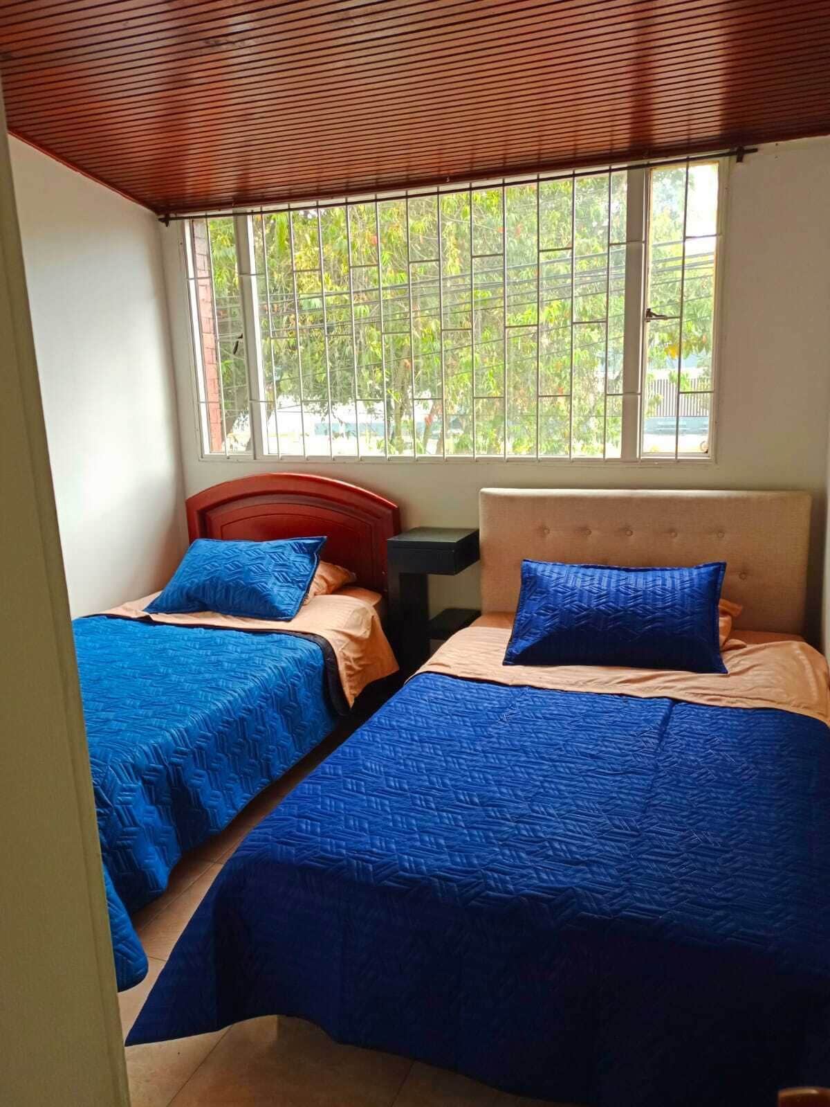 A room with two beds, each with blue bedding and a wooden headboard. There is a window with metal bars, and a view of greenery outside.