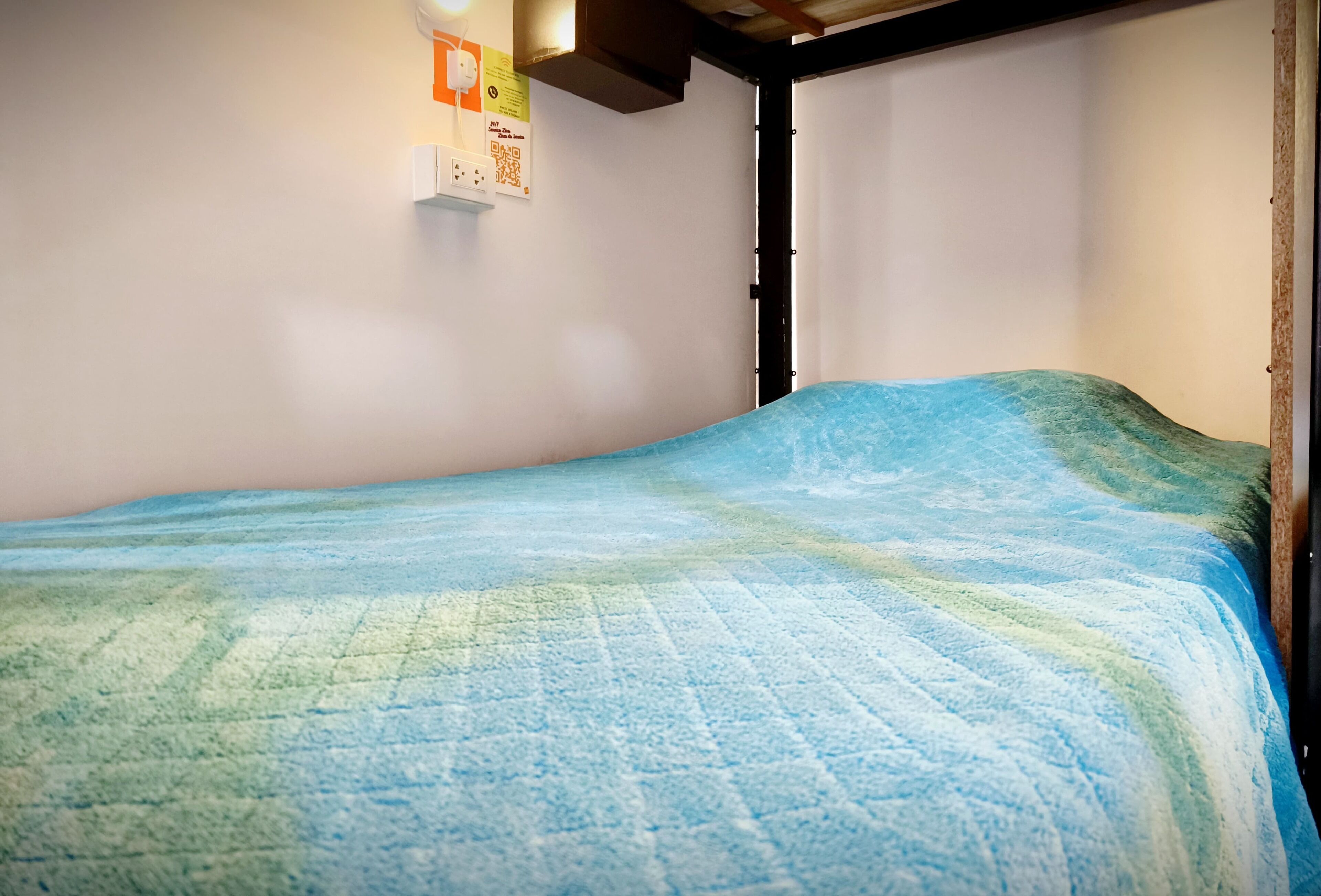 A single bed with a blue and green quilt in a room with white walls and a wooden ceiling.