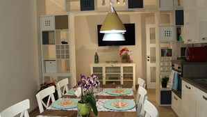 Un coin repas avec une table dressée pour quatre personnes, entouré de chaises blanches, un téléviseur fixé au mur et un espace cuisine avec des placards et un four à micro-ondes.