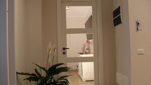 Un couloir avec une plante en pot, une porte dotée d’un miroir et un aperçu d’une pièce où l’on aperçoit un lit et une table de chevet.