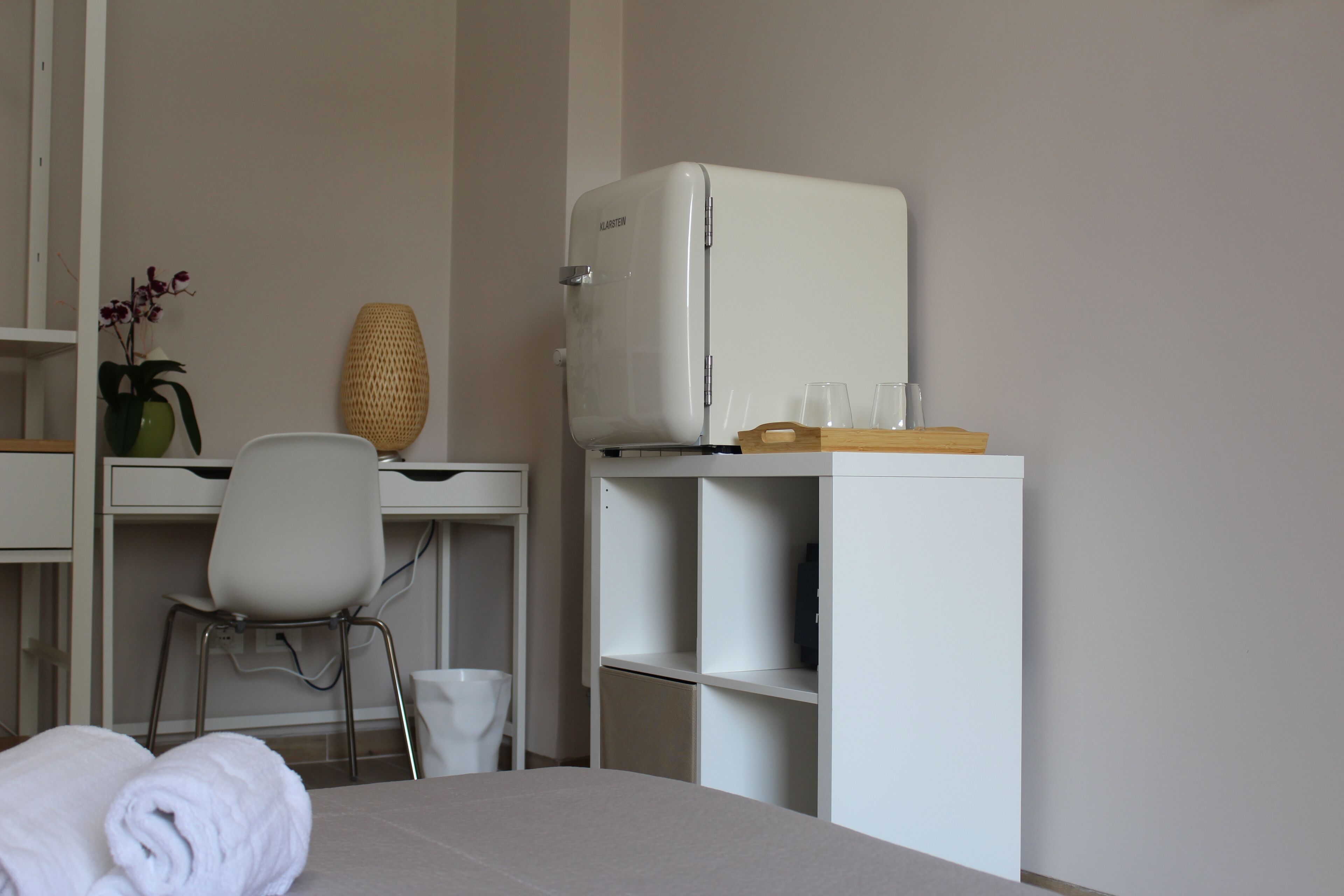 Ein Hotelzimmer mit einem Bett, einem Schreibtisch mit Stuhl, einem kleinen Kühlschrank und einem Regal mit Gläsern.