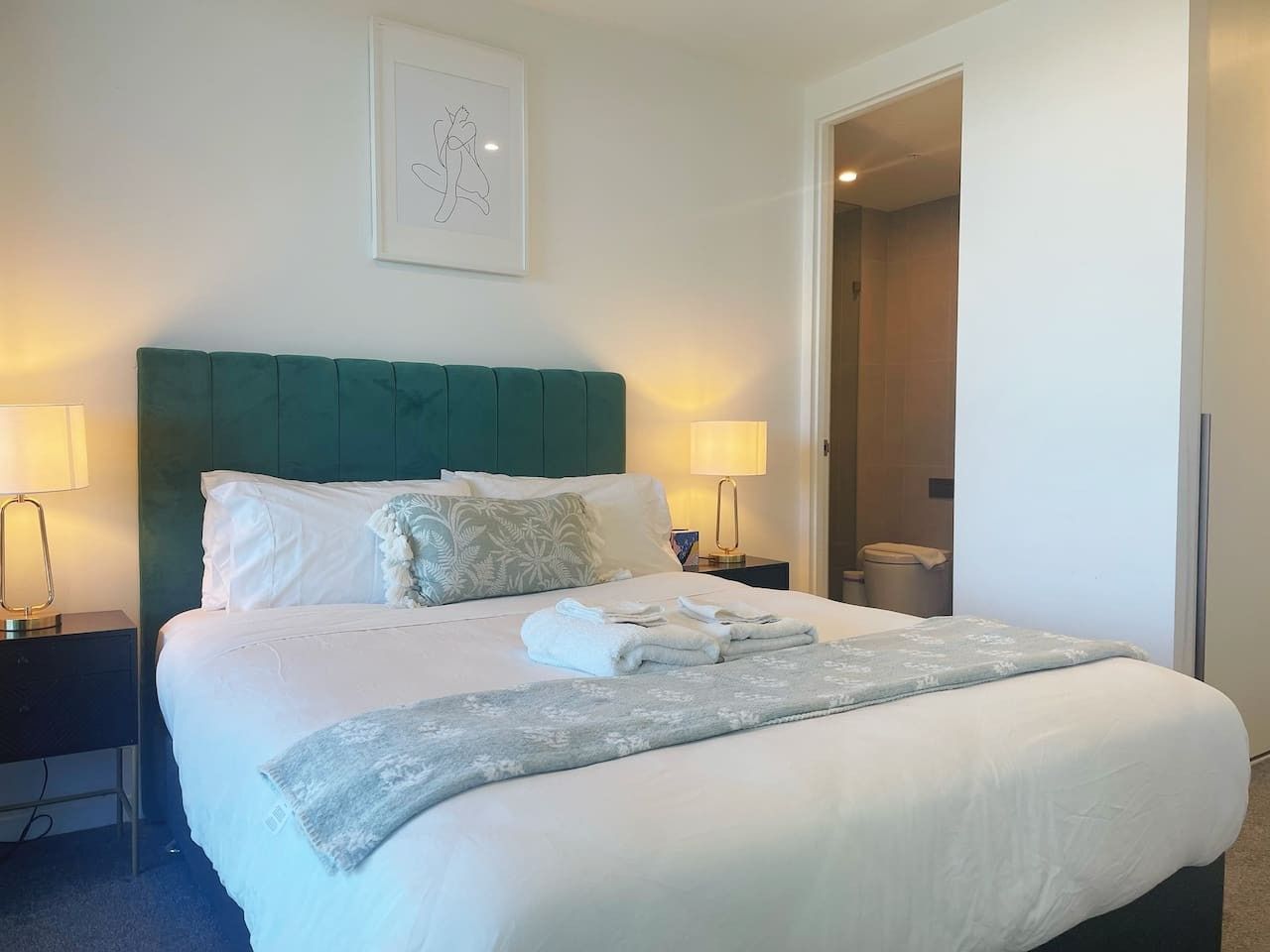 A neatly made bed with white linens, a teal headboard, and a framed artwork above it.