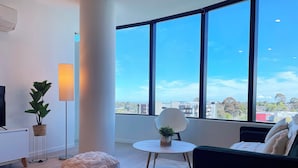 A modern living room with a large window, a flat-screen TV, a floor lamp, a potted plant, a round ottoman, and a sofa with cushions. - Panoramic Views! Spacious 2 Bed, 2 Bath Luxury Apartment w/ Free Parking (Glen Waverley)