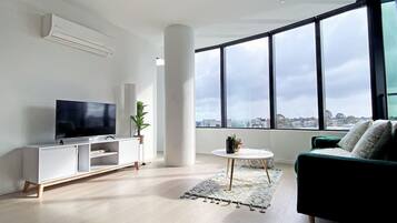 A modern living room with a flat-screen TV, white entertainment unit, green sofa, and a large window offering a city view.