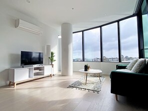A modern living room with a flat-screen TV, white entertainment unit, green sofa, and a large window offering a city view.