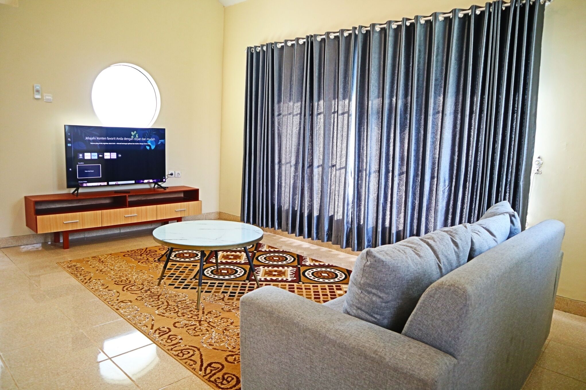 A living room with a sofa, a TV on a wooden stand, a patterned rug, and a curtain.