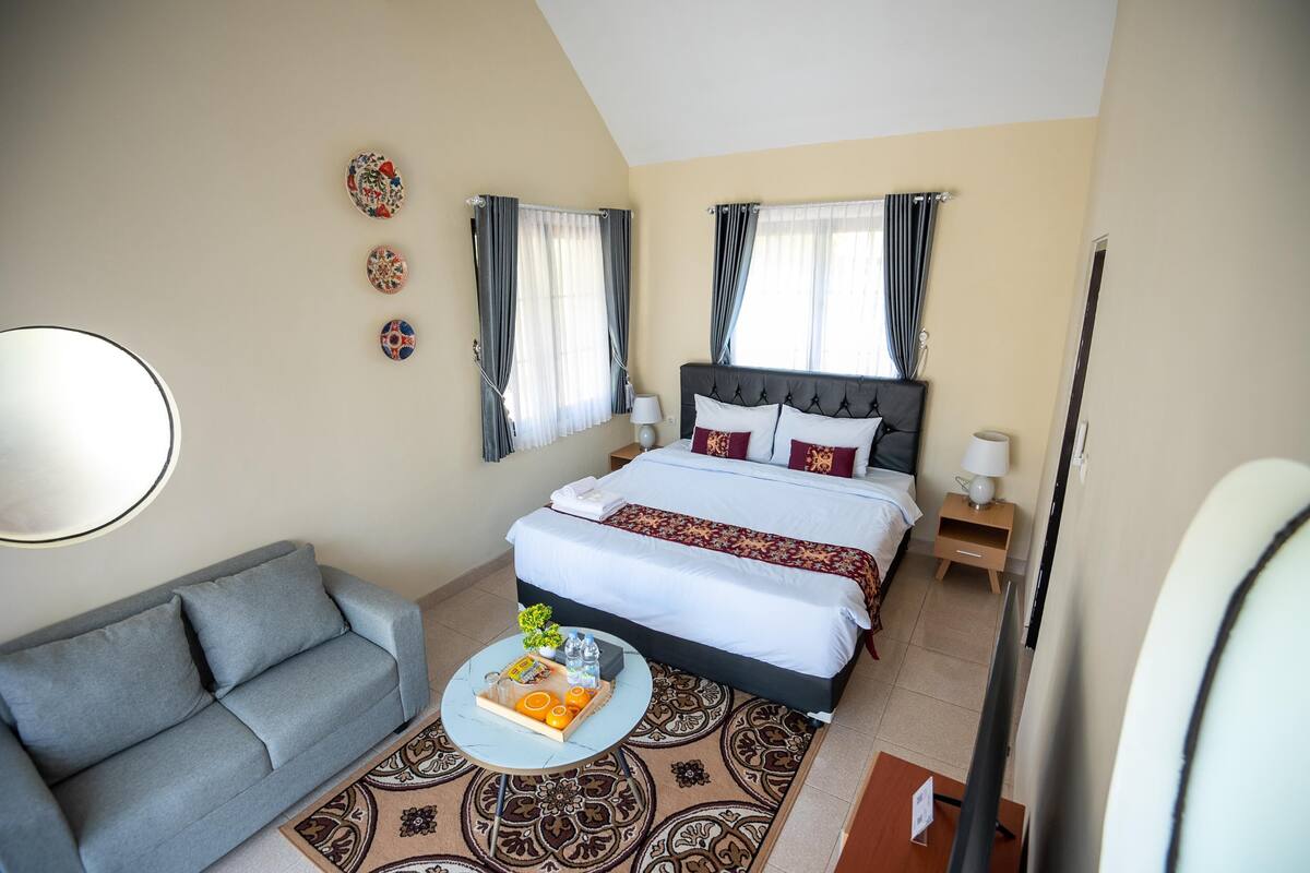 Una habitación de hotel con cama, sofá, mesa de centro con frutas y un libro, mesita de noche con lámpara, y tres adornos en la pared.