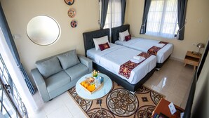 Quarto de hotel com sofá, duas camas, mesa de centro redonda com frutas e bebidas, e tapete estampado.