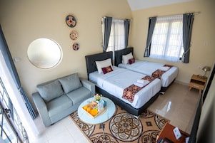 Una habitación de hotel con un sofá, dos camas, una mesita redonda con fruta y bebidas, y una alfombra estampada.