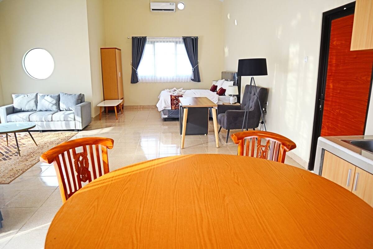 Habitación de hotel con mesa de comedor, sillas, sofá, cama, armario, televisor y ventana con cortinas.