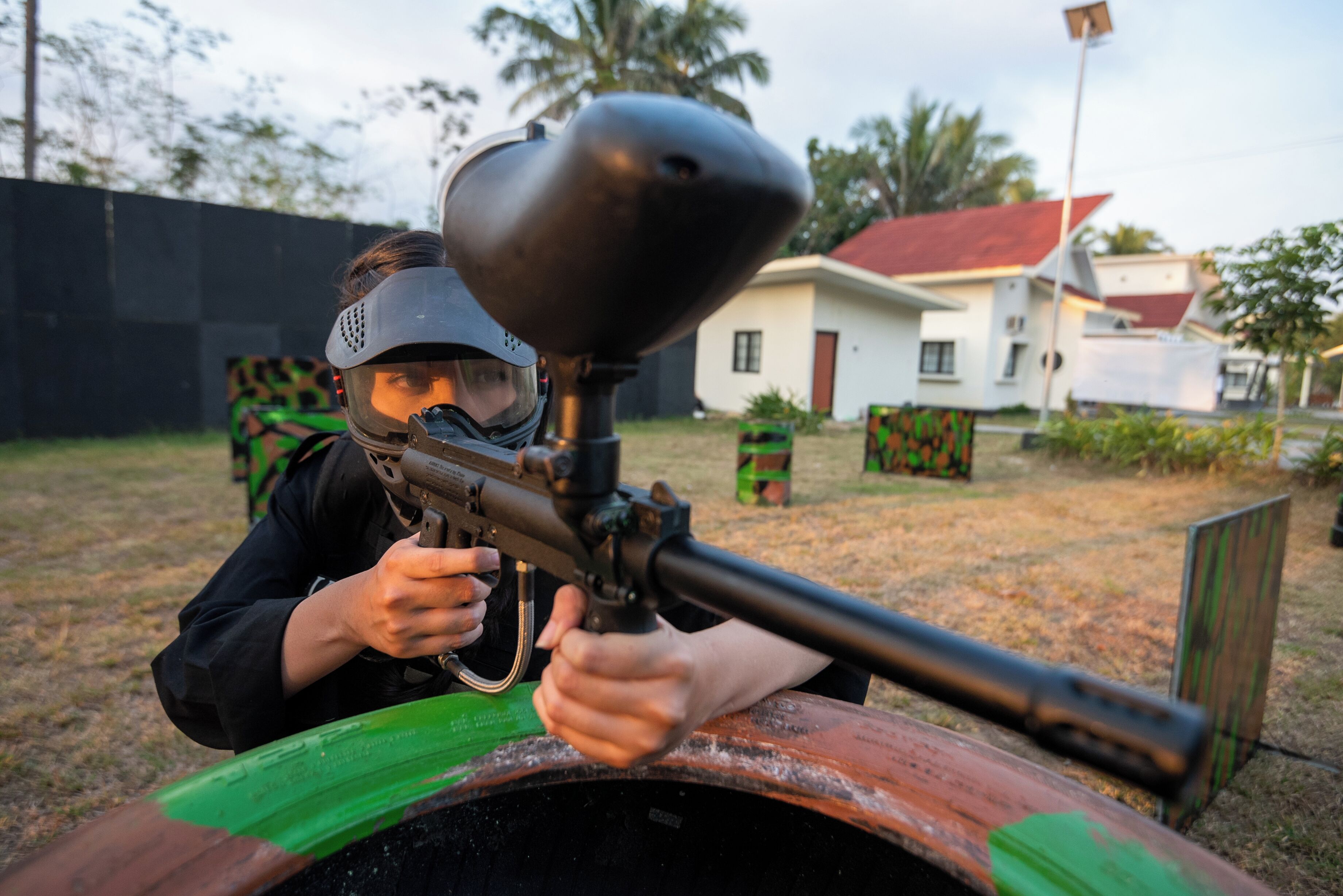 A person aiming a paintball gun from behind a wooden barrel.