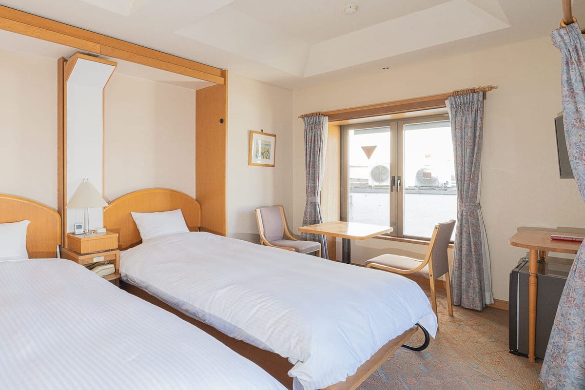 Hotelkamer met twee enkele bedden, een kleine tafel, een stoel, een bureau en een raam met gordijnen.