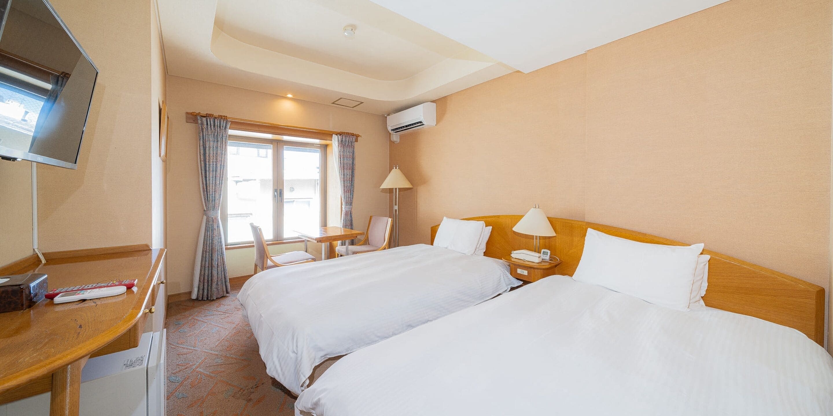 Ein Hotelzimmer mit zwei Betten, einem Schreibtisch mit Fernseher, einem Tisch mit Stühlen und einem Fenster mit Vorhängen.