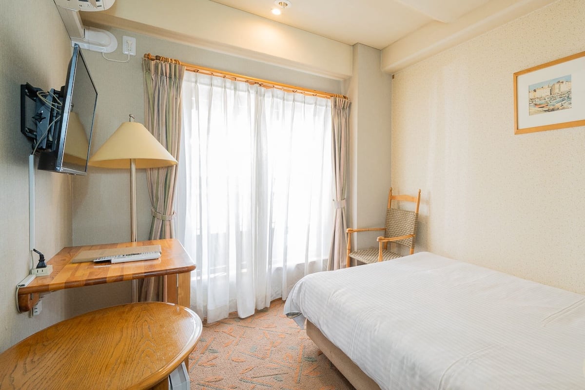 Een hotelkamer met een bed, een bureau, een stoel, een lamp en een raam met gordijnen.