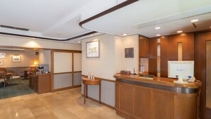 Een hotel receptie met een houten balie, een zithoek met stoelen en een tafel, ingelijste foto's aan de muur en een computerscherm op de balie.