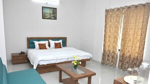 A bedroom with a bed, a wooden bedside table with a vase of flowers, a wooden nightstand, and a blue sofa. - Hotel Pride Arena (Bhopal)