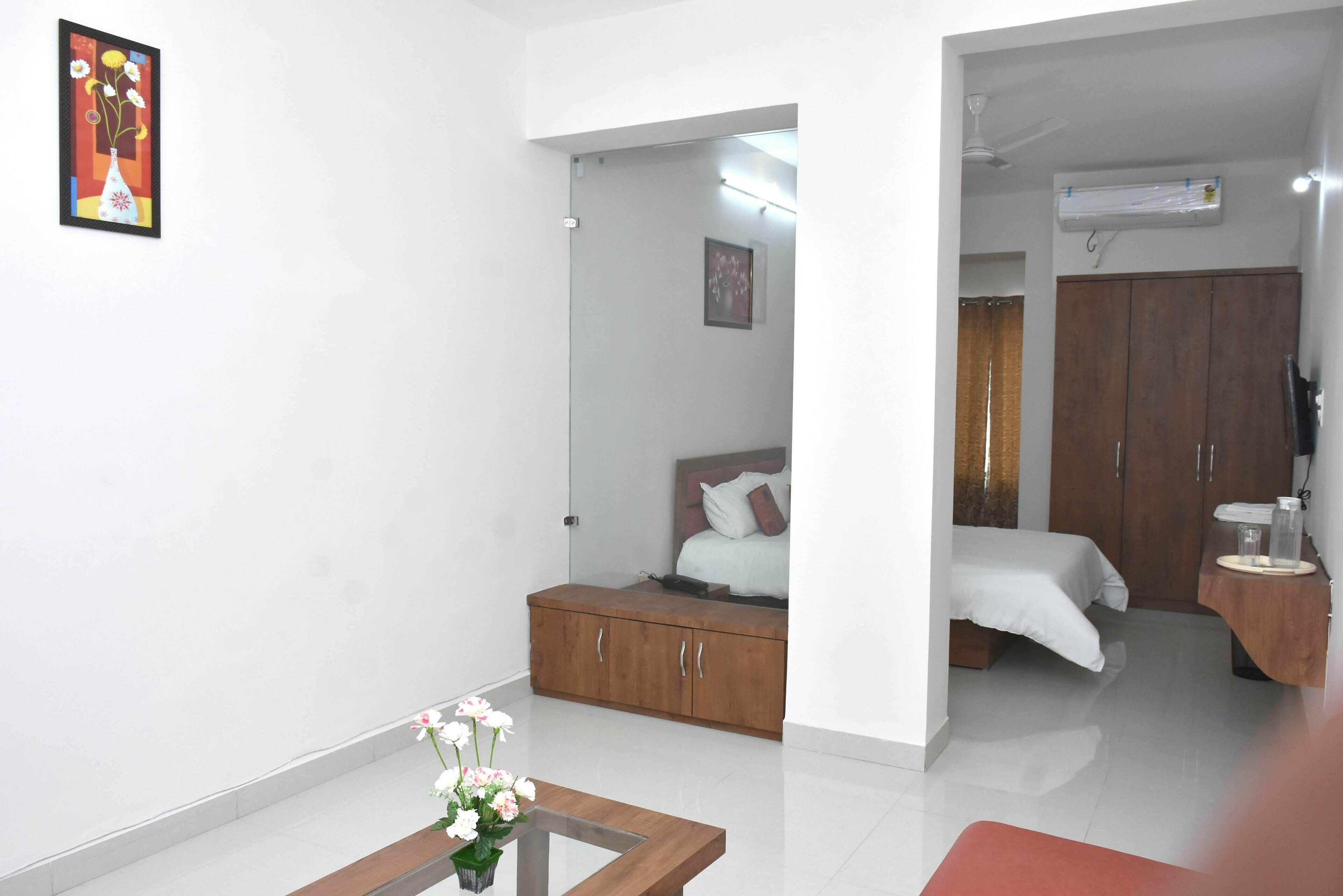 A hotel room with a bed, a wooden wardrobe, a small table with a vase of flowers, and a framed picture on the wall.