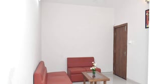 A small, modern living room with a red sofa, a wooden coffee table with a vase of flowers, and a wooden door. - Hotel Pride Arena (Bhopal)