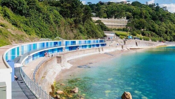 A curved swimming pool with clear blue water, surrounded by a sandy beach and lush greenery. - Spacious Apartment, 1 Minute from Seafront (Paignton)