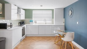 A modern kitchen with white cabinets, a microwave, and a sink. There is a dining table with chairs and a blue wall in the background. - Spacious Apartment, 1 Minute from Seafront (Paignton)
