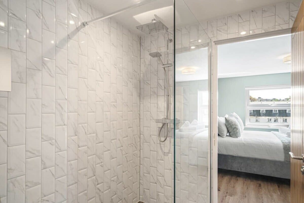 Une salle de bain moderne dotée d’une douche à parois de verre, de murs à motifs de marbre et offrant une vue sur une chambre avec un lit et une fenêtre.