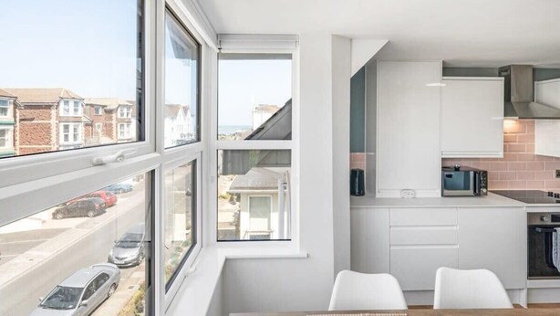 Una cucina moderna con mobili bianchi, un forno a microonde e un fornello. Ci sono due sedie bianche a un tavolo, e un'ampia finestra offre vista sulla strada e sulle case vicine.