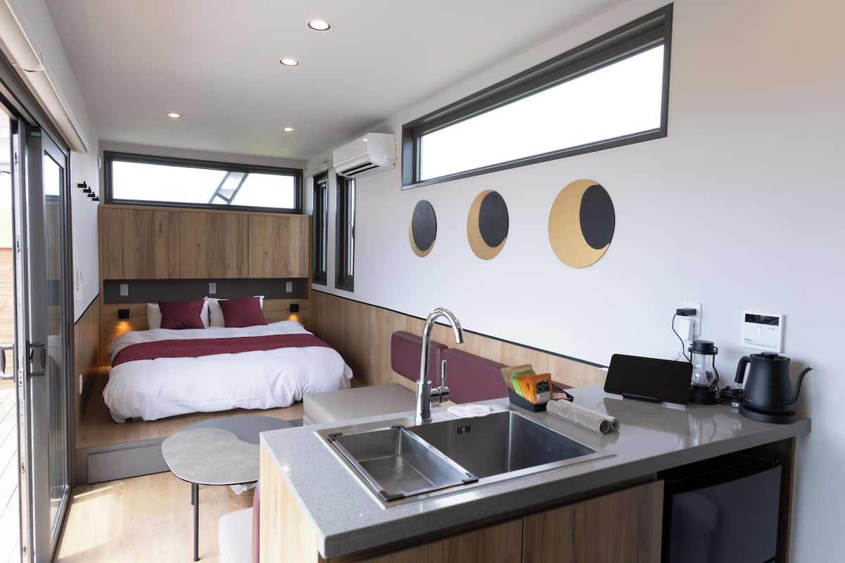 A modern hotel room with a bed, a small sitting area, a kitchenette with a sink, and a large window.