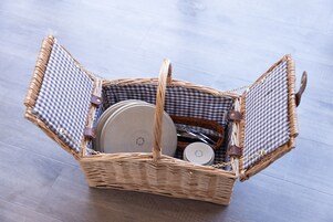 Un cesto di vimini foderato di tessuto a quadretti, contenente un piatto bianco, un piccolo contenitore e una bottiglia.