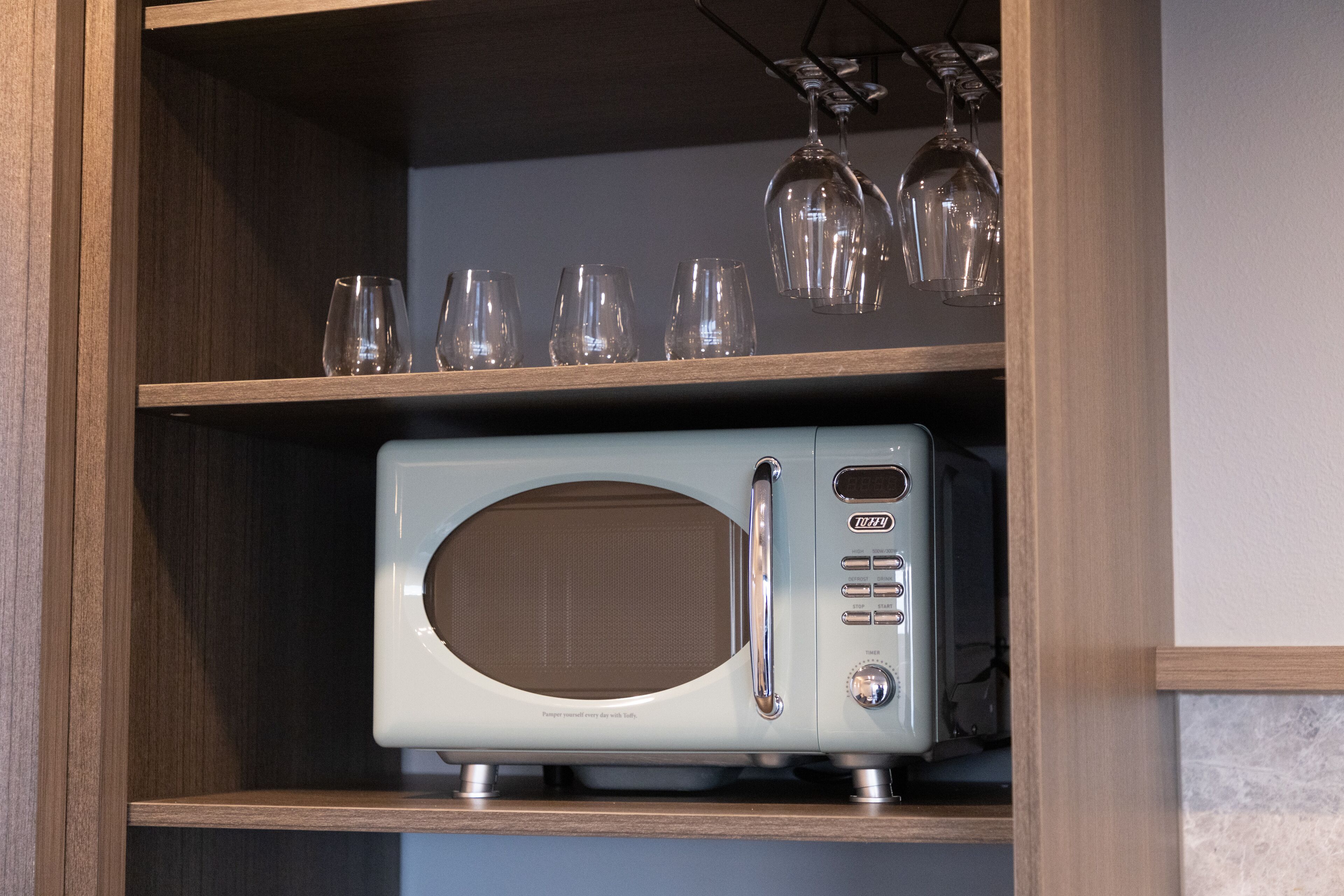 A kitchen with a microwave on the countertop and wine glasses on the shelf above.