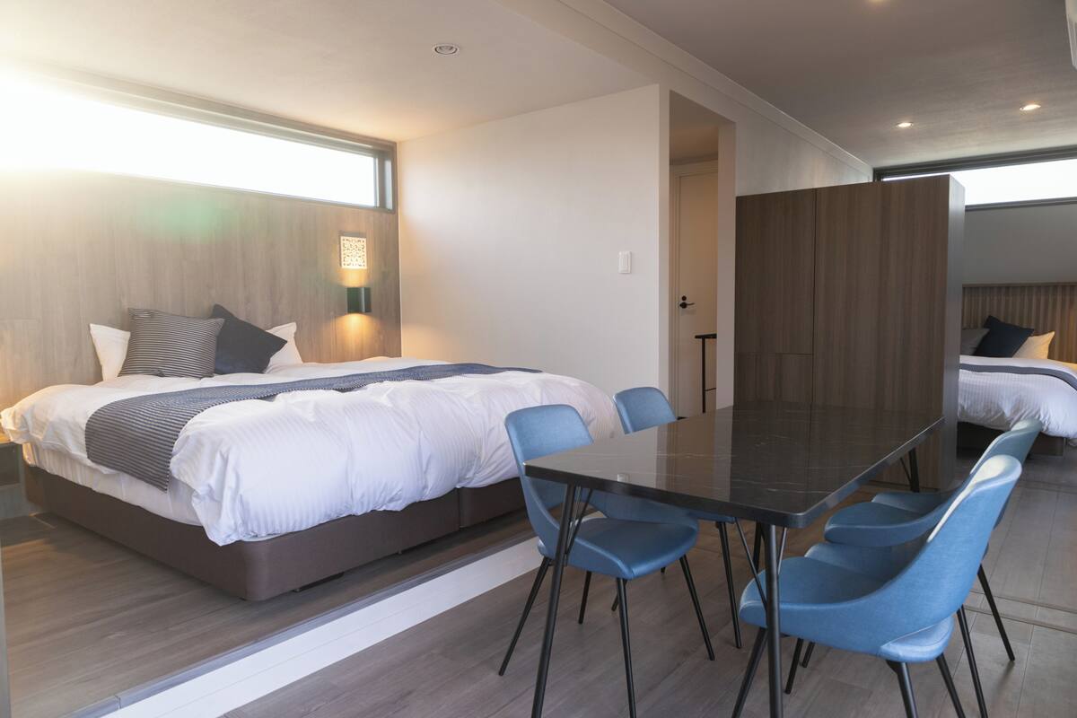 A modern hotel room with a large bed, a dining table, and blue chairs.
