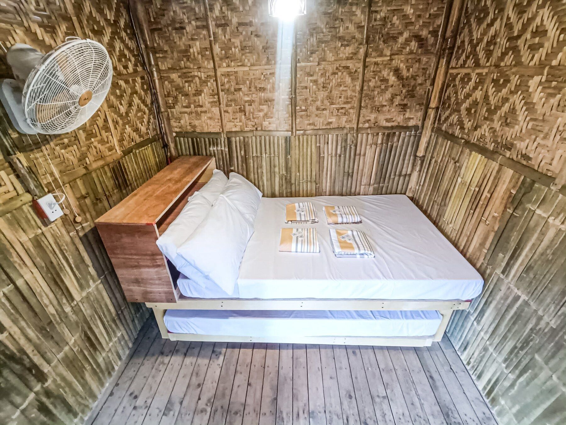 A compact room with a bed, a wooden bench, and a wall-mounted fan.