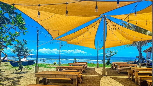 Ein Sitzbereich am Strand mit Holzbanken unter großen, gelben Sonnensegeln, mit Blick aufs Meer.
