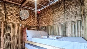 Ein Einzelbett mit Holzrahmen und blauer Matratze, ein Wandventilator und eine gewebte Bambuswand.