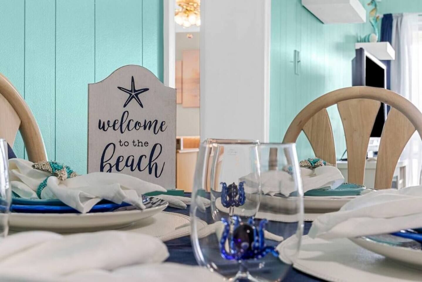 A dining table set with plates, glasses, and a decorative item, with a sign reading 'welcome to the beach' in the background.