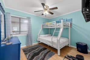 A bunk bed with a ceiling fan, a desk with a bird sculpture, and a trash bin.