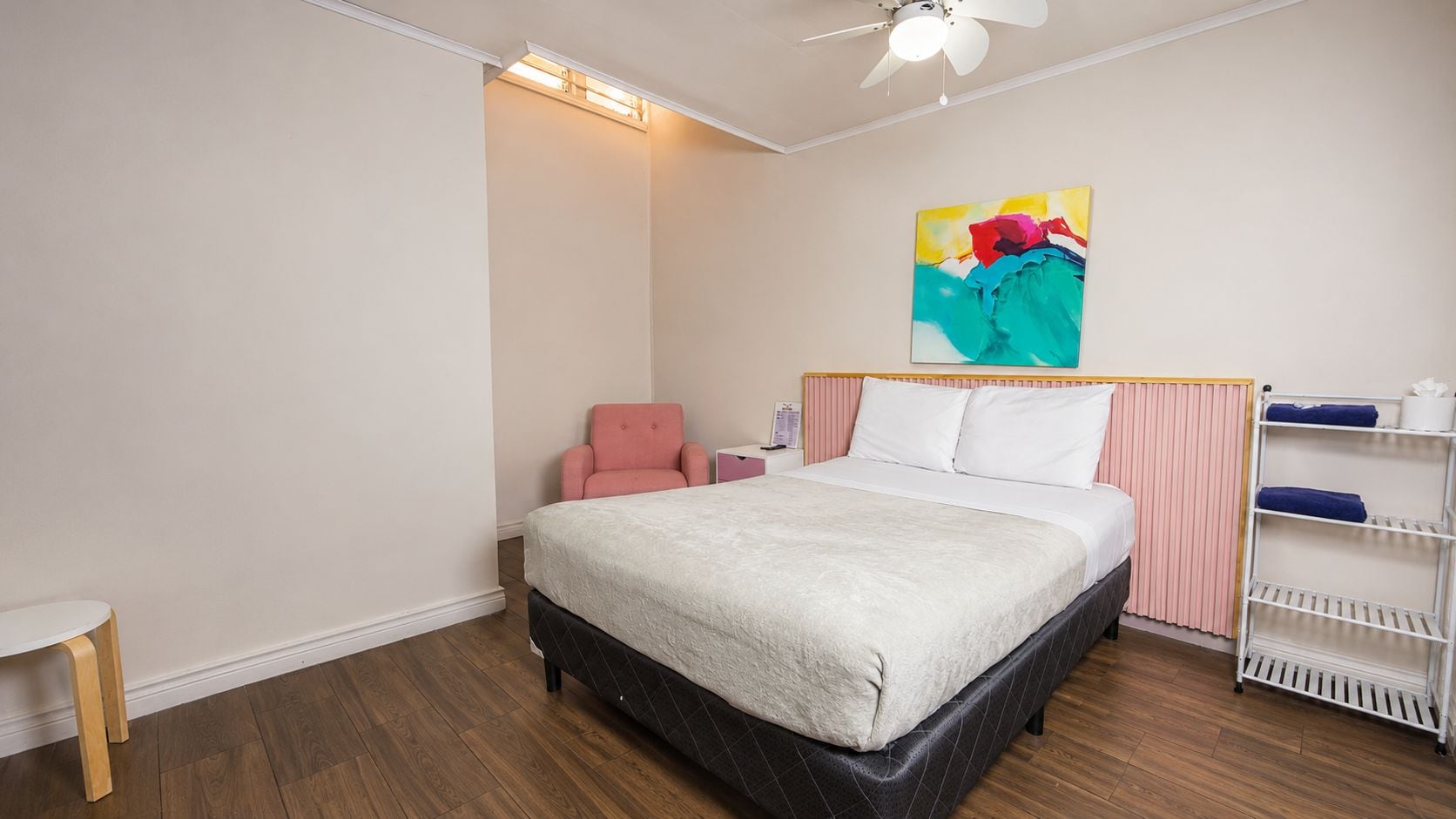 Une chambre d’hôtel avec un lit, une chaise, une étagère et un ventilateur de plafond.