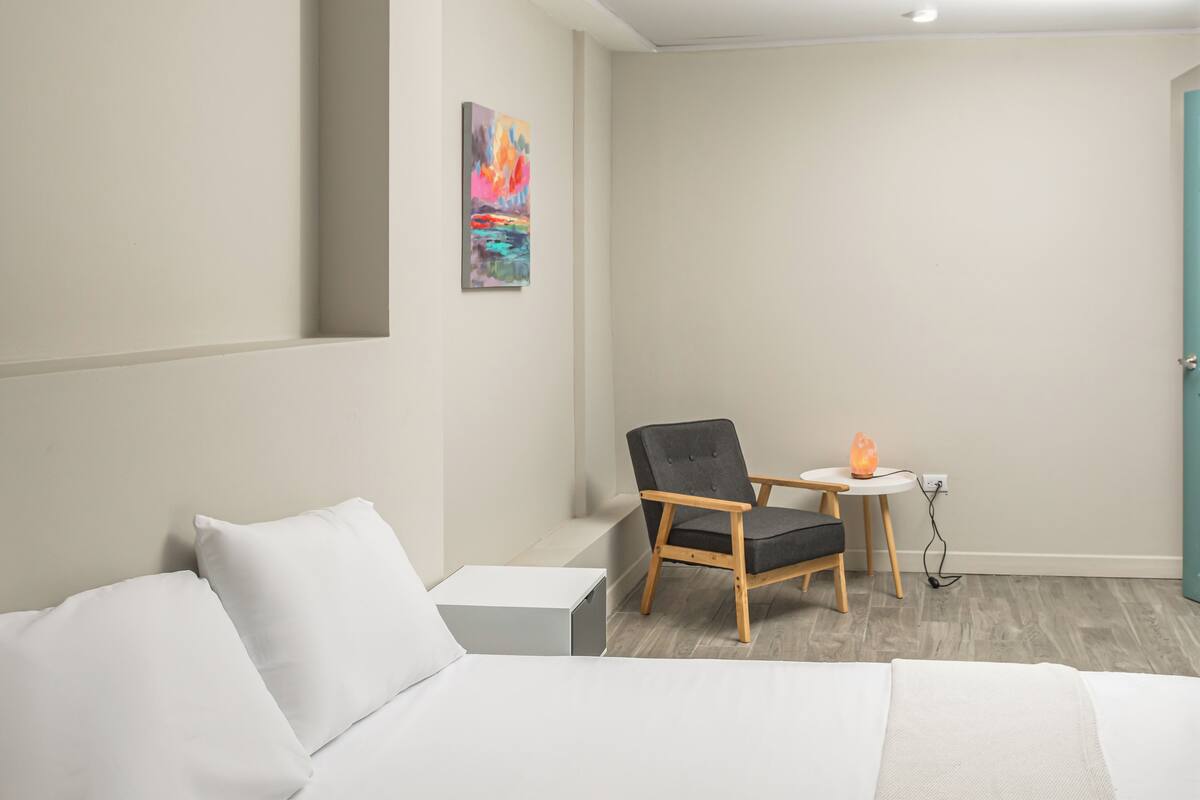 Et moderne hotelv ærelse med en seng, en stol, et lille bord med en varmeovn og et billede på væggen.