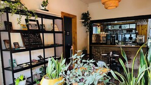 A cozy cafe interior with a wooden counter, black metal shelving with plants, and a hanging light fixture. - Otoya1907 Hotel (San José)