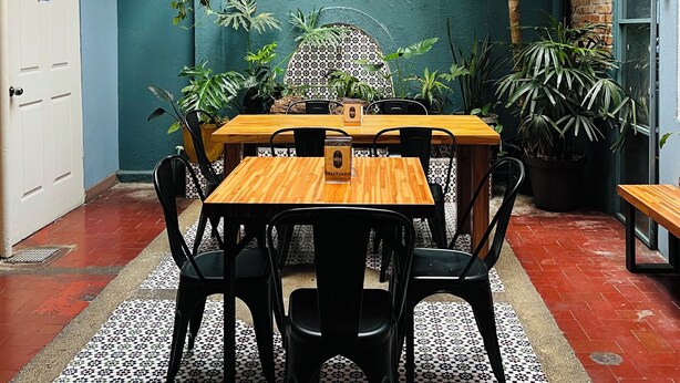 A dining area with a wooden table and chairs, hanging wicker lamps, and potted plants. - Otoya1907 Hotel (San José)