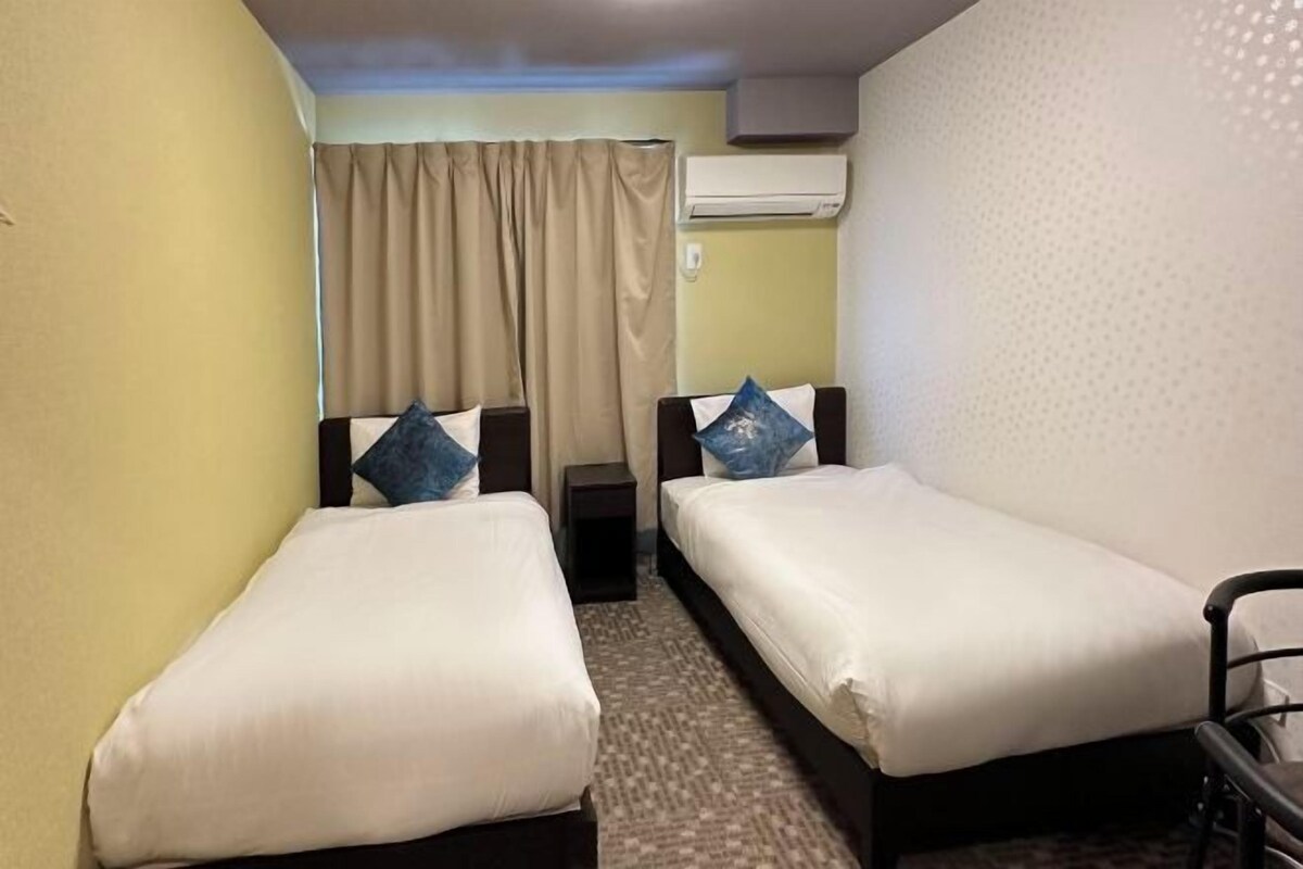 A hotel room with two single beds, a small table, an air conditioning unit, and a window with curtains.