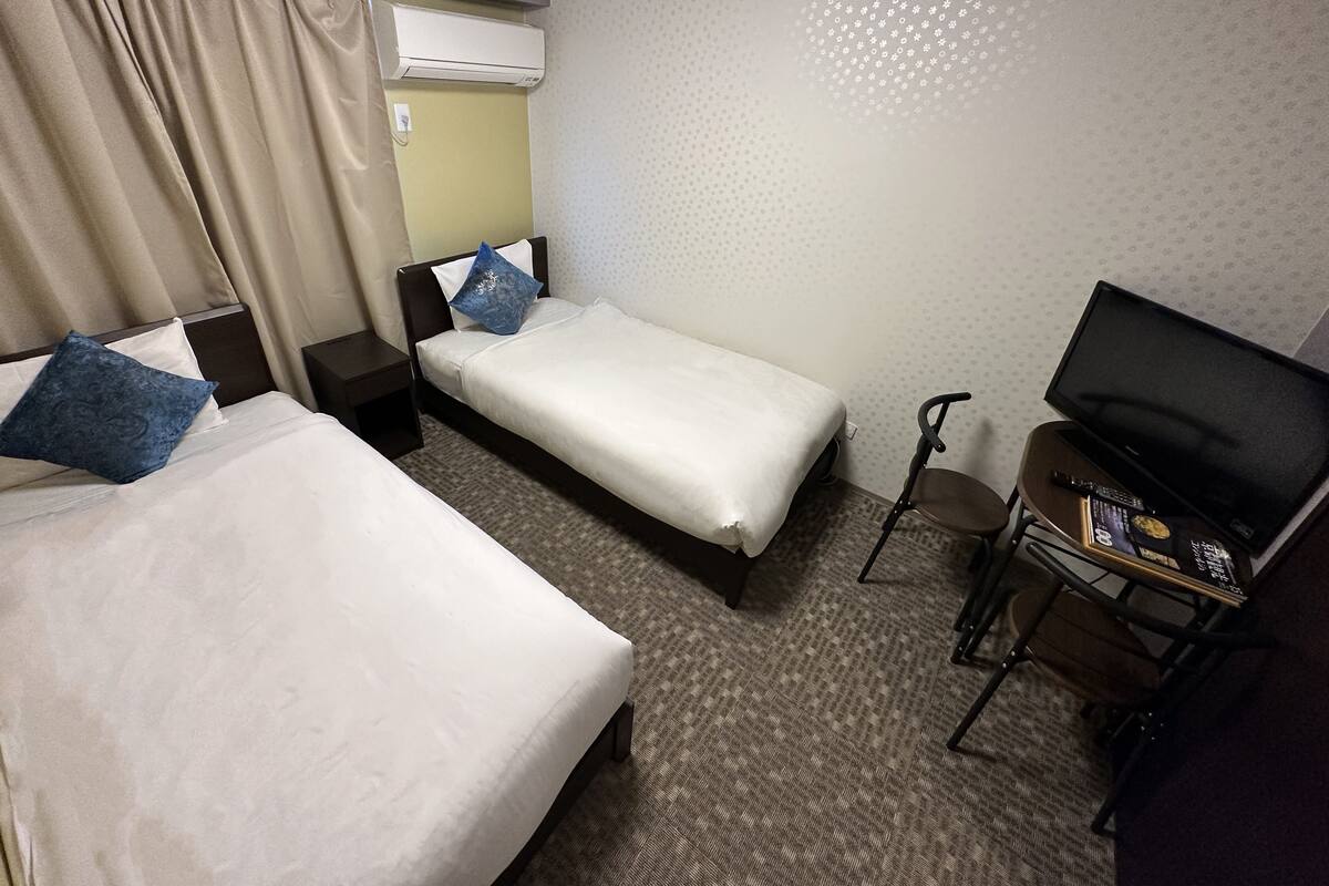 A hotel room with two single beds, a TV, a desk, and an air conditioning unit.