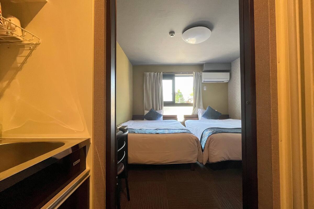 A hotel room with two beds, a window with curtains, an air conditioning unit, and a wooden floor.