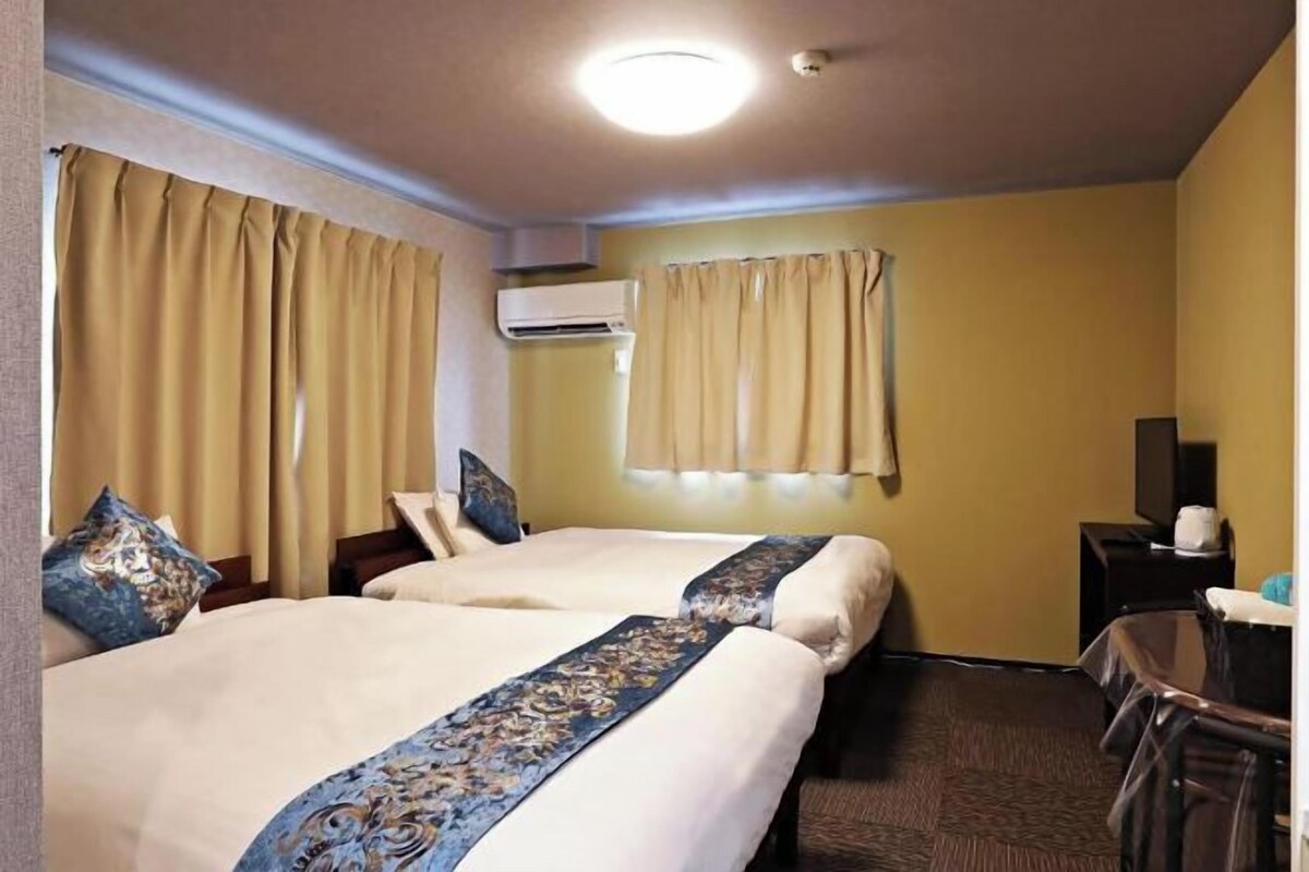 A hotel room with two beds, beige curtains, an air conditioning unit, a desk with a chair, and a television.