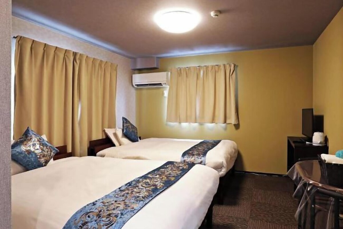 Une chambre d’hôtel avec deux lits, des rideaux beiges, un climatiseur, un bureau avec une chaise et un téléviseur.