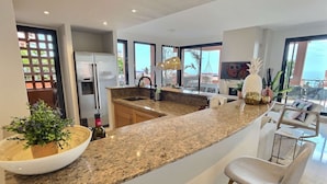 Uma cozinha moderna com bancadas de granito, uma ilha central, banquetas e uma grande janela com vista para o oceano.