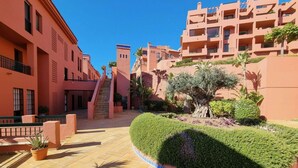 Um pátio com uma escadaria central, rodeado por edifícios com varandas e um jardim bem cuidado com diversas plantas.