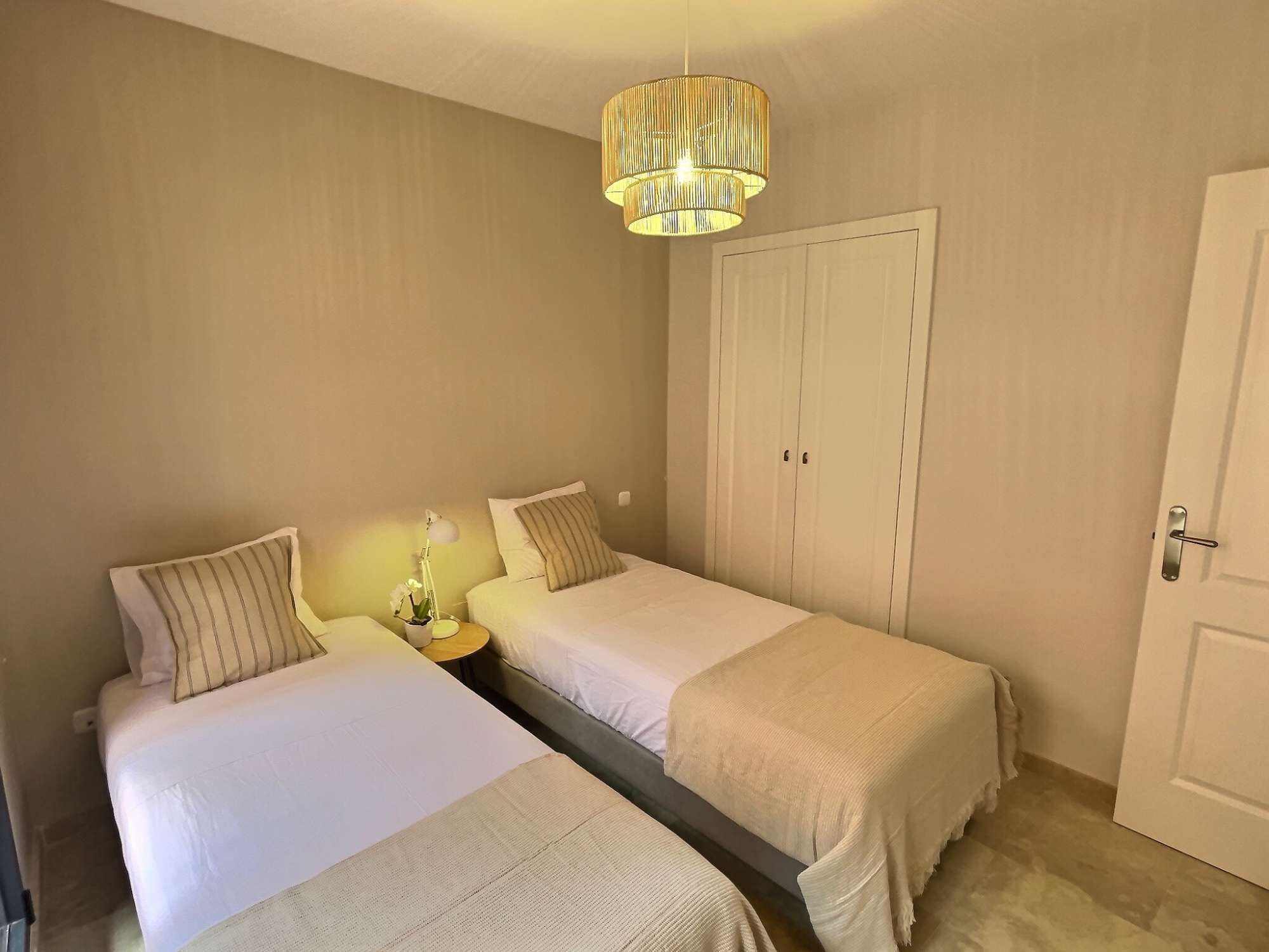A bedroom with two beds, a wardrobe, a nightstand with a lamp, and a hanging light fixture.